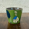 Handmade ceramic mug with green glaze, blue-roofed house, tree, and clouds design, perfect for coffee or tea.