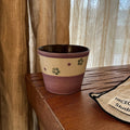 Handmade ceramic mug with green floral pattern, purple rim, and speckled beige band, on wooden table near curtain