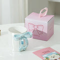 White ceramic mug with blue bow handle beside pink gift box, perfect for tea or coffee lovers